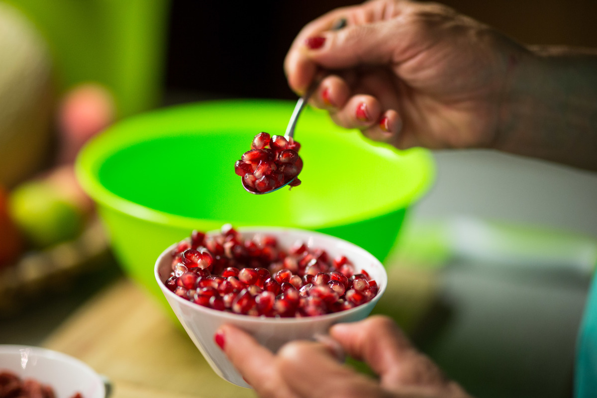 A woman takes a teaspoon of pomegranate seeds from a bowl she holds in her left hand